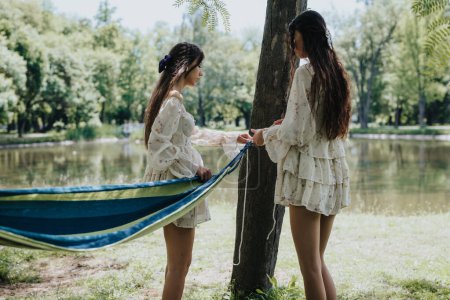 A peaceful scene depicting two friends putting up a blue hammock beside a calm lake in a lush park, reflecting a serene day outdoors.