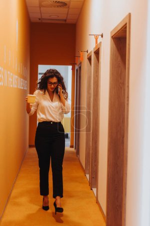 A young businesswoman negotiates projects and enjoys tea in a modern co-working space. She walks confidently while talking on the phone.