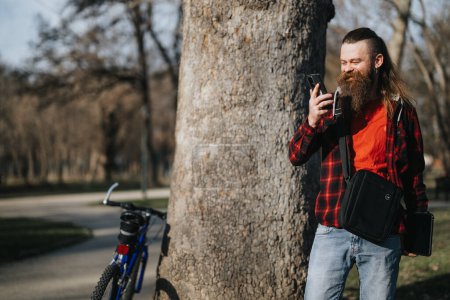 Bärtiger Geschäftsmann in Freizeitkleidung, der mit Smartphone und Fahrrad im Stadtpark arbeitet, was für Flexibilität bei der Arbeit aus der Ferne steht.