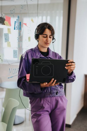 Konzentrierte indische Geschäftsfrau mit einem digitalen Tablet in einem modernen Büroumfeld, mit klebrigen Notizen und Zahlen im Hintergrund.