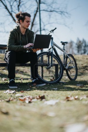 Ein gutaussehender Mann genießt einen entspannten Arbeitstag im Freien auf seinem Laptop, sitzend neben seinem Fahrrad in einem sonnigen Park.