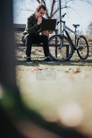 Ein gut aussehender junger Mann sitzt an einem sonnigen Tag mit seinem Laptop auf einer Parkbank, neben ihm sein Fahrrad, das eine perfekte Mischung aus Arbeit und Freizeit darstellt..