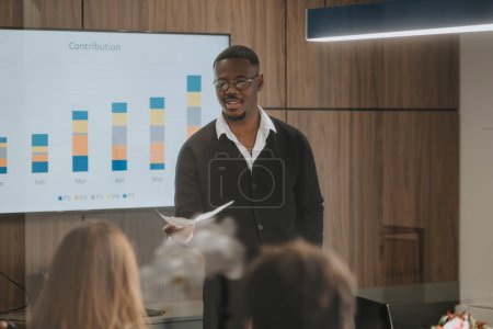 Confident young entrepreneur presents financial statistics to colleagues in a well-lit meeting room, emphasizing company growth and collaboration.