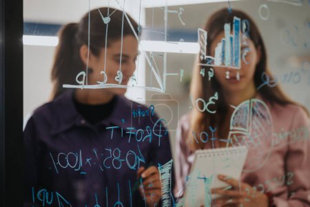 Deux femmes d'affaires participent à une séance stratégique, interprétant et discutant des statistiques complexes écrites sur un mur de verre transparent à l'aide de marqueurs et de notes collantes.