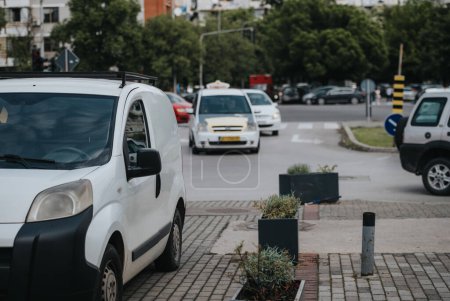 Städtische Straßenszene mit einem geparkten weißen Lieferwagen, belebten Kreuzungen mit anderen Fahrzeugen, Grünflächen und Gebäuden im Hintergrund. Autos stecken im Stau fest.