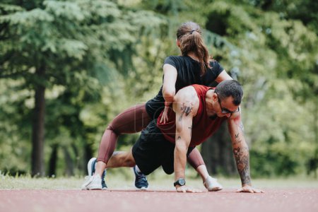 An energetic photo capturing a fitness duo in a park a woman assists a man doing push-ups, signifying teamwork and strength in fitness training.