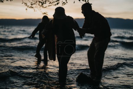 Dans une scène dramatique au bord du lac, un homme soutient les femmes qui naviguent sur de fortes vagues sous un ciel sombre et atmosphérique.