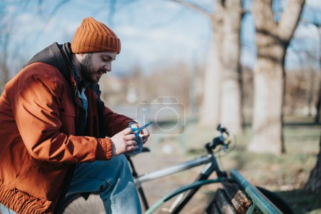Hombre enfocado con barba en un cálido gorro naranja y chaqueta roja usando un teléfono inteligente, sentado junto a su bicicleta en un parque soleado.