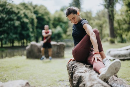 Focused young woman wearing sportswear stretches after a workout on a log in a lush green park, with a blurred male athlete resting in the background.