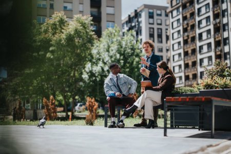 Three diverse startup colleagues engaged in a business conversation outdoors near modern city buildings, expressing ideas and teamwork.