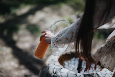 Una joven con el pelo castaño largo lleva un vestido floral ligero mientras rellena su botella de agua naranja en un día soleado..