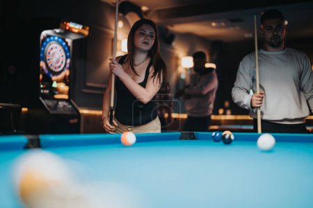 Eine fokussierte junge Frau spielt mit einem männlichen Partner in einer schwach beleuchteten Bar Billard. Freunde und Unterhaltung im Hintergrund verstärken die ungezwungene, lustige Atmosphäre der Szene.