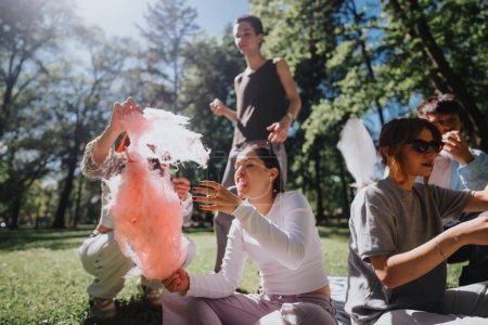 Eine Gruppe junger Freunde entspannt sich und genießt Zuckerwatte an einem sonnigen Tag in einem üppig grünen Park, der Freiheit und Freude verkörpert.