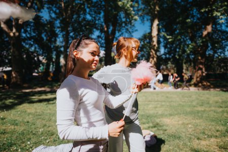 Dos mujeres jóvenes sonriendo y disfrutando de dulces de algodón rosa al aire libre en un parque vibrante en un día soleado.