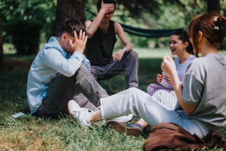 Scène en plein air avec un jeune homme frustré tenant la tête dans un parc, avec deux femmes et un autre homme en arrière-plan riant et parlant.