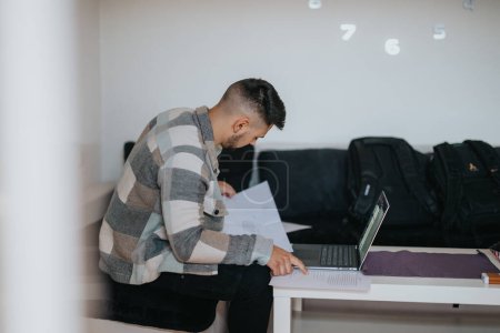 Un jeune adulte concentré examine la paperasse tout en utilisant un ordinateur portable sur un bureau blanc à la maison, dépeignant la concentration et le dévouement.
