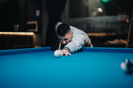Ein junger Mann konzentriert sich intensiv, während er in einer stilvollen, modernen Poolhalle bei einem Wettkampfspiel einen Schuss abgibt.