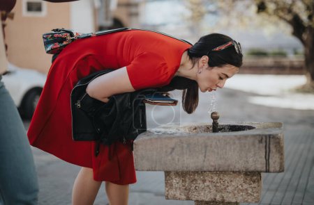 Refreshed woman in vibrant red enjoying a drink from an urban water fountain on a warm, sunny day outdoors.