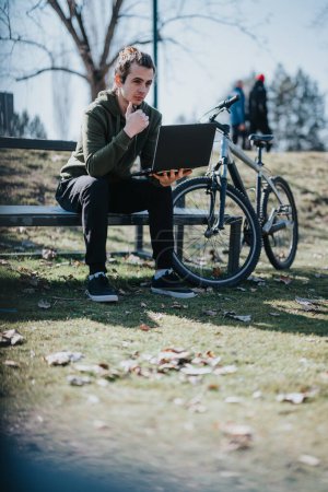 Lässiger junger Mann genießt einen sonnigen Tag im Park, indem er an seinem Laptop arbeitet, auf einer Bank neben seinem Fahrrad sitzt und die moderne Freiberuflichkeit verkörpert.