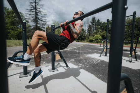 A fit man with tattoos exercises using outdoor gym equipment in a park on a clear, sunny day, showcasing health and active lifestyle.