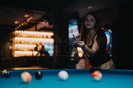 Mujer joven enfocada con palo de indicadores apuntando a bolas de billar en una mesa de billar en un bar con poca luz.