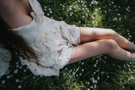 An overhead view of a young girl lying in a flower print dress, enjoying a peaceful day in a grassy park with blooming daisies.