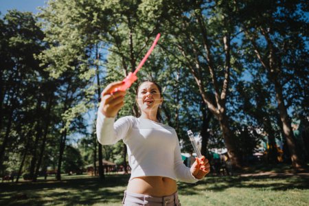A young woman joyfully blows soap bubbles on a sunny day at the park, surrounded by lush green trees and the warm sunlight.