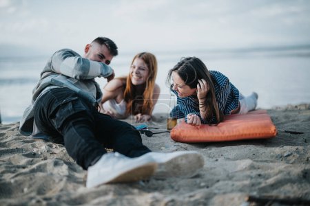 Three young adults share laughter and stories, conveying joy and relaxation during a beach hangout at dusk, showcasing a sense of friendship and leisure.