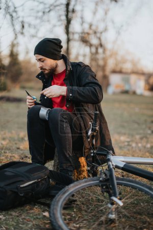 Konzentrierter junger Mann hält beim Radfahren inne und sitzt mit Klebeband neben seinem Fahrrad in einem ruhigen Park, als die Sonne untergeht.