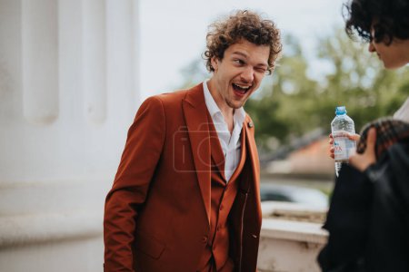 Joyful man in an orange suit winking at the camera while talking with a friend outdoors, expressing happiness and excitement.