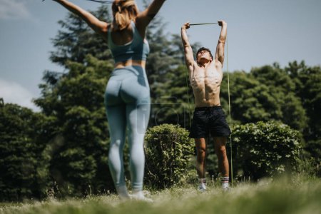 Young fit man and woman working out with resistance bands in a sunny park. Outdoor fitness routine highlighting strength and healthy lifestyle.