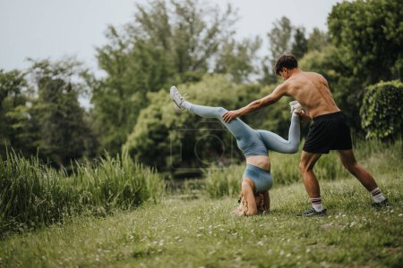 Fit couple practicing handstand during workout in the park. Man assisting woman with balance exercise on grassy field surrounded by greenery.