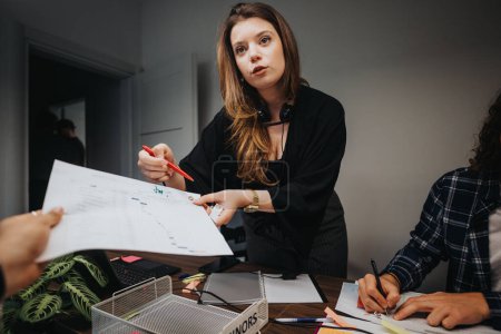 A professional young woman leads a business discussion with diverse colleagues, highlighting charts and engaging in teamwork at a small, organized office.