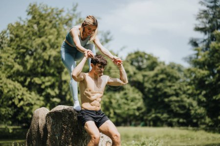 Two fit friends doing a workout outdoors, balancing and supporting each other on a rock amidst lush greenery on a sunny day.
