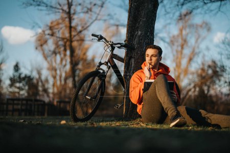 A thoughtful young man rests near his bike in a tranquil park setting as the sun sets, exuding a sense of leisure and freedom.