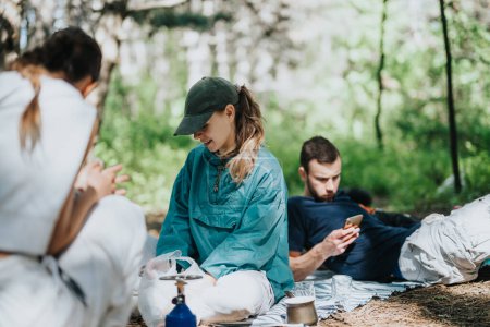 Group of friends relaxing during a camping trip in a forest, one engaged with a smart phone and others enjoying nature.
