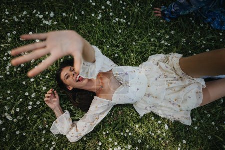 Fröhliches junges Mädchen in einem floralen Kleid, das auf einem mit Gänseblümchen gefüllten Gras in einem Park liegt und spielerisch in die Kamera greift.