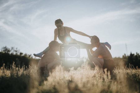 Flexible girl performing front split exercise with the support of her male friends on their shoulders in a field during sunset, showcasing strength, balance, and teamwork.