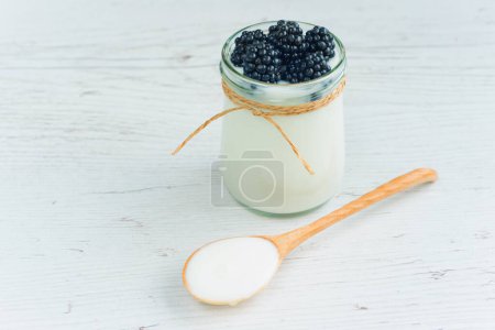 Foto de Yogurt in small jar with fresh berries. Blackberries in yogurt. Yogurt is in jar on the wooden table. Wooden spoon full of yogurt lies on the table - Imagen libre de derechos