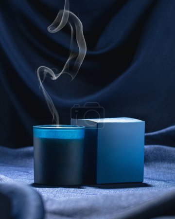 Foto de Una vela azul perfumada con una caja azul fuma sobre un telón de fondo de tela azul oscuro - Imagen libre de derechos