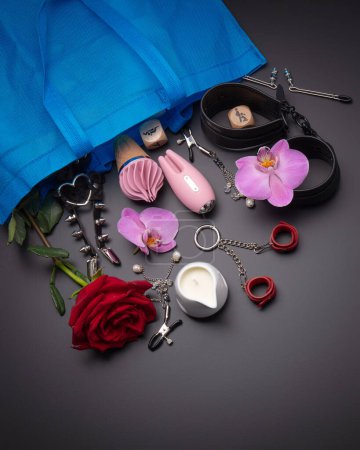Photo for Collection of intimate pleasure devices featuring pink vibrators and  elegant bondage accessories, accompanied by delicate orchids and red rose for touch of romance on dark backdrop - Royalty Free Image