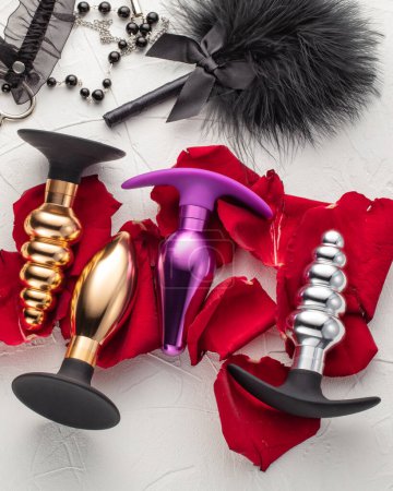 Photo for Collection of anal plugs of different shapes and colors presented on bed of red flower petals against white textured background with elegant black intimate accessories. Sex toys for adults - Royalty Free Image