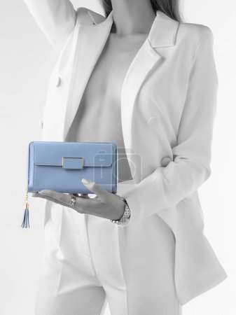 Monochrome image of woman in white formal suit with unbuttoned jacket exposing body, holding elegant blue leather clutch with gold details and tassel. Stylized photo with partial desaturation effect