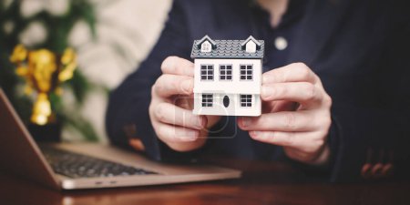 Photo for Real estate agent holding house model in front of laptop. Real estate - Royalty Free Image