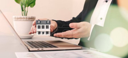 Photo for Real estate agent holding house model in front of laptop. Real estate business - Royalty Free Image