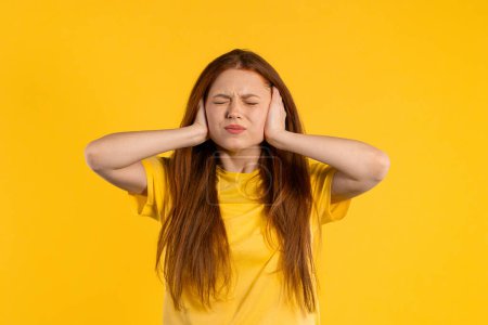 Irritierte Ingwerfrau mit verdeckten Ohren auf gelbem Hintergrund. Teenager verbieten störende Geräusche, Schreie oder Geräusche. . Hochwertiges Foto