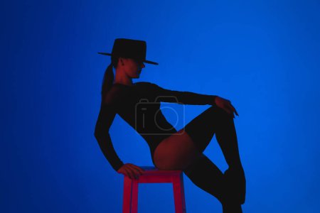 Photo for Professional ballet dancer in hat sitting on high chair on multi-colored background under violet neon light. Sensual sexy ballerina dancing with her legs in stockings. Femme fatale outfit. High photo - Royalty Free Image