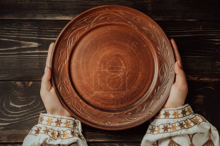 Manos femeninas con plato de barro ucraniano vacío sobre mesa de madera. Mujer con camisa bordada tradicional. Vista superior. Vajilla nacional con adorno hecho a mano. Foto de alta calidad