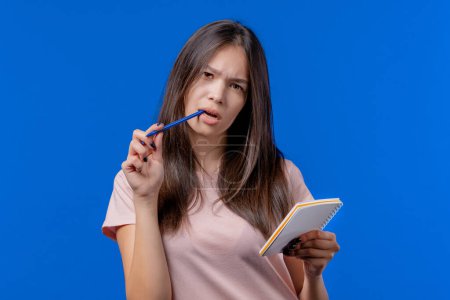 Foto de Young woman writing notes in planner with pen. Student girl thinking about future plans and to-do list in notebook for week, month, year. Keeping personal diary on blue background. High quality photo - Imagen libre de derechos