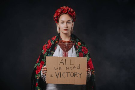 Foto de Ukrainian woman in traditional costume with cardboard All we need is victory on blue background. Ukraine win war. Democracy, liberty, demonstration, russian aggresion concept. High quality photo - Imagen libre de derechos